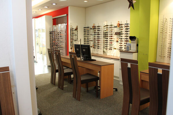 Modernes Optikergeschäft mit Beratungstisch, Computer und vielen Brillen an der Wand.